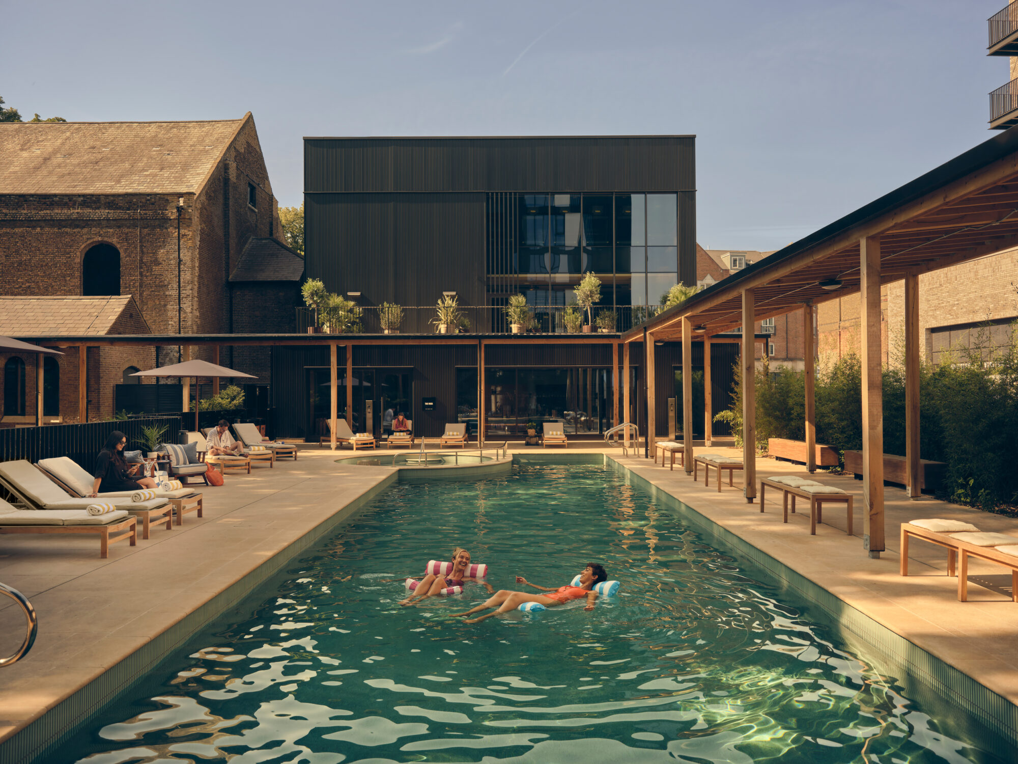 The outside area of The Brentford Project, known as the Resident's Cub, with people in the pool, an outdoor area with chairs and an oak timber-covered walkway.