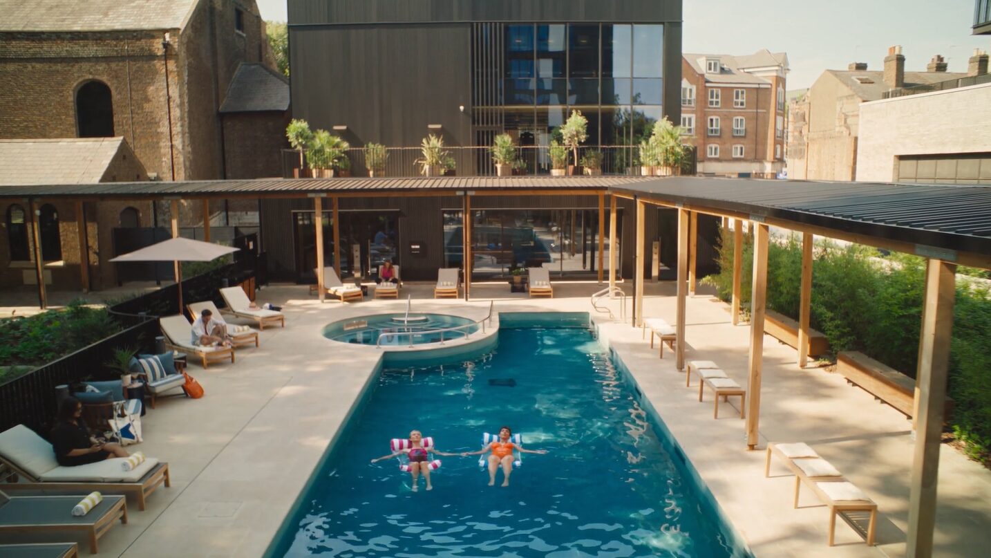 The outside area of The Brentford Project, known as the Resident's Cub, with people in the pool, the outdoor area with chairs and an oak timber-covered walkway covering the outside area around the pool.